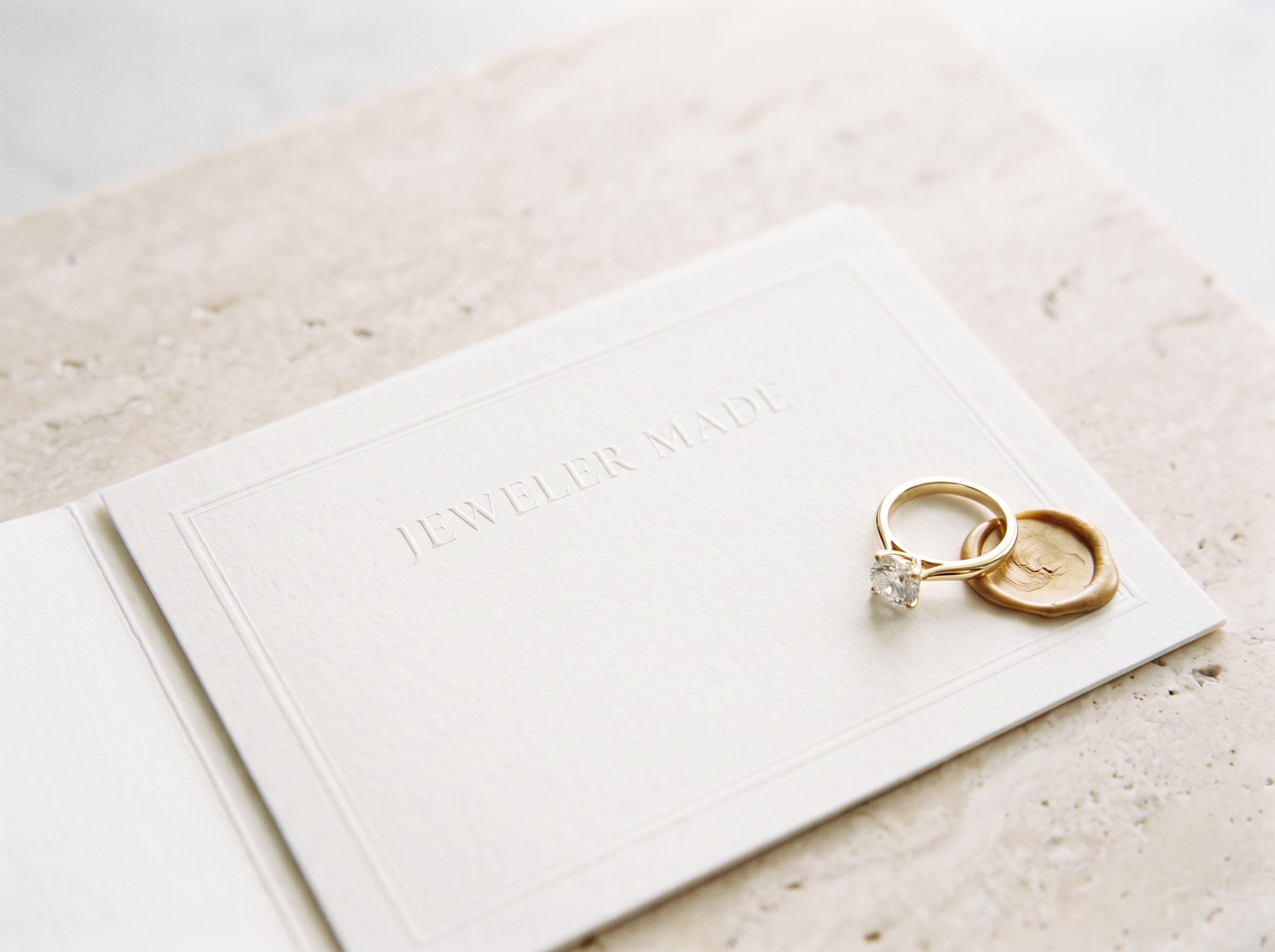 An ivory certificate blind-embossed with the Jeweler Made wordmark, a gold solitaire ring and a gold wax seal resting on its corner